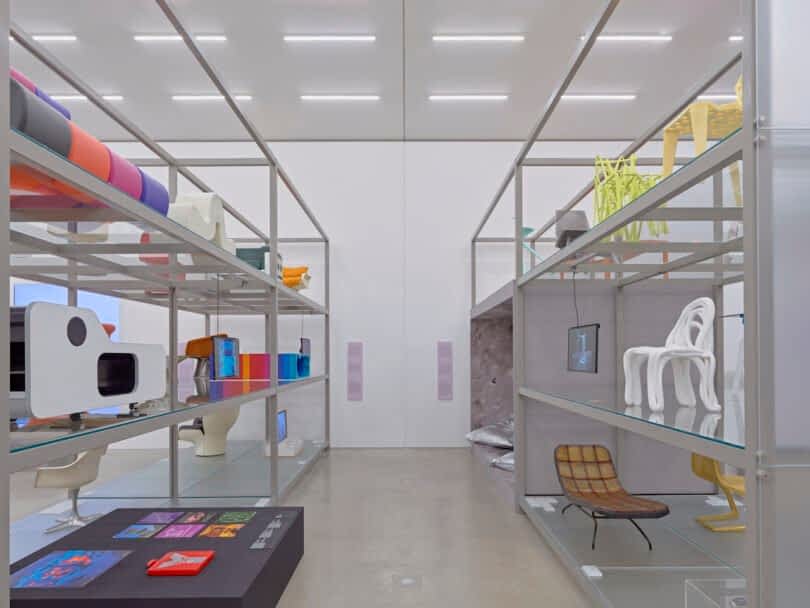A modern display room with various art and design pieces on metal shelves, featuring colorful and uniquely shaped items including chairs, screens, and abstract sculptures