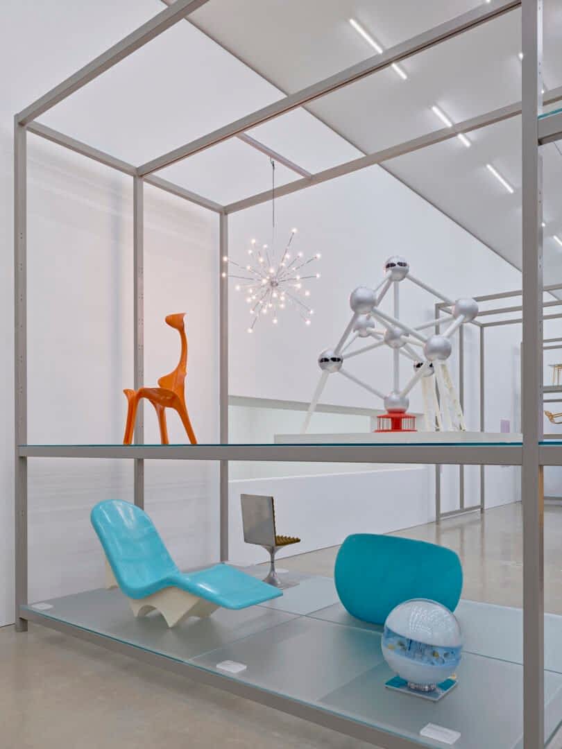 Display of various modern design objects set on metal shelving, including a blue chair, orange giraffe figure, small model of an atom-like structure, and a spherical lamp