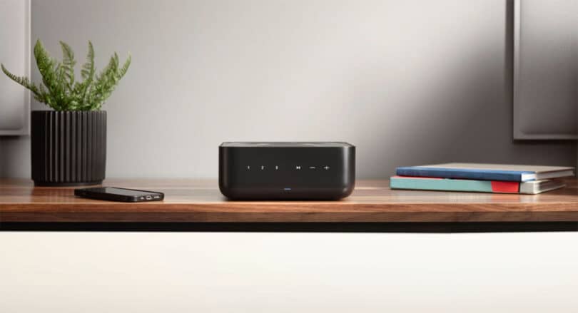 A sleek Denon Home Amp wireless speaker with touch controls sits on a wooden desk, accompanied by a smartphone, a potted plant, and a stack of books.