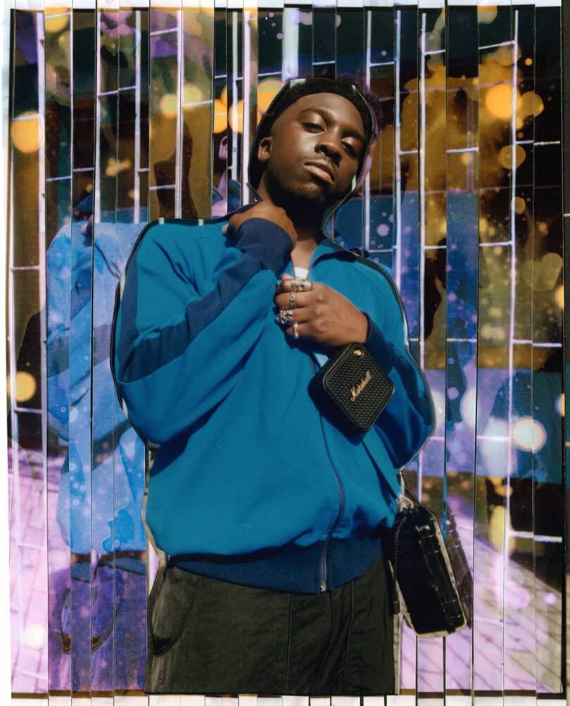 A man wearing a blue jacket stands in front of a vibrant, abstract background with vertical lines and colorful light patterns, holding the collar of his jacket with one hand and a small Marshall bag with the other.