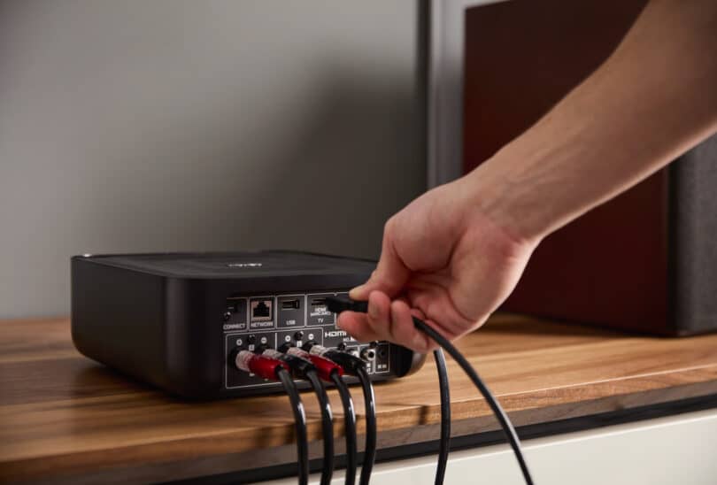 A person plugs an HDMI cable into the back of the Denon Home Amp placed on a wooden surface, surrounded by other connected cables.