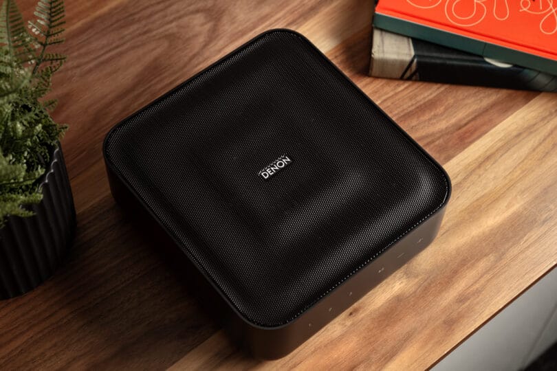 A black Denon Home Amp sits on a wooden surface beside some books and a potted plant.