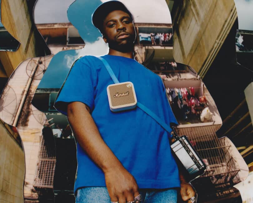 A person wearing a blue shirt, blue hat, and shoulder bag stands in front of a cityscape background. They have a small speaker hanging across their chest.