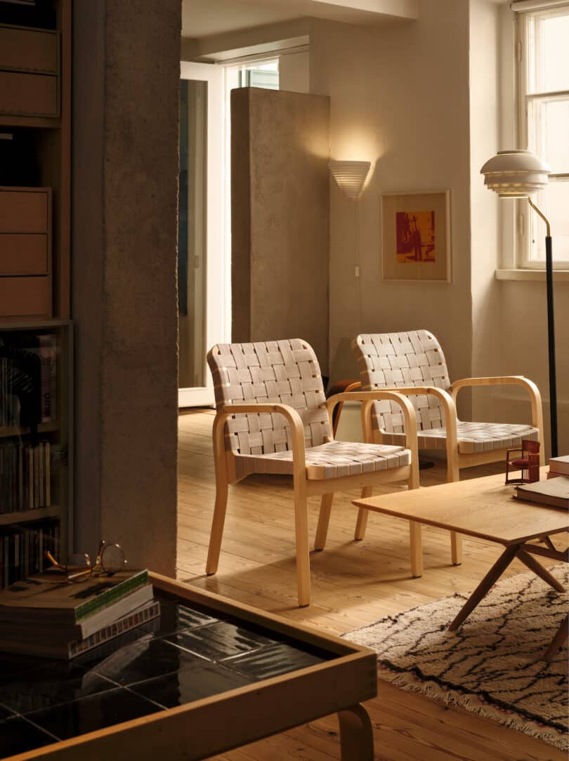 Two modern beige armchairs with wooden frames in a warmly lit living room, accompanied by a wooden coffee table. A lamp is mounted on the wall, and a large window lets in natural light