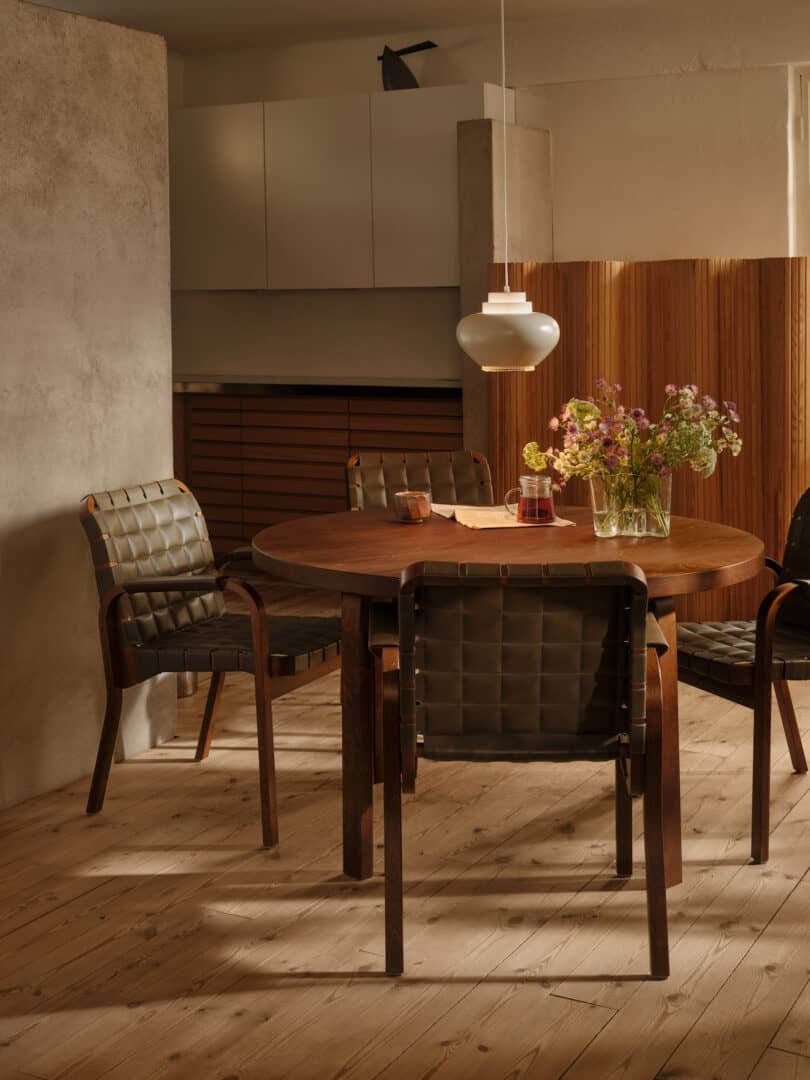 A rustic dining area with a round wooden table, woven leather chairs, a hanging pendant light, and a vase of flowers. The background features a mix of wooden and concrete elements