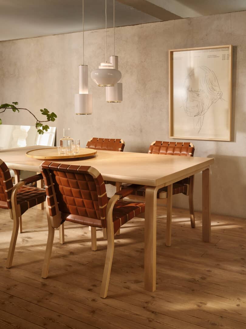 A beige-walled dining room with a wooden table, four brown woven chairs, three pendant lights, a framed sketch, a potted plant, and a tray with glasses and a water carafe