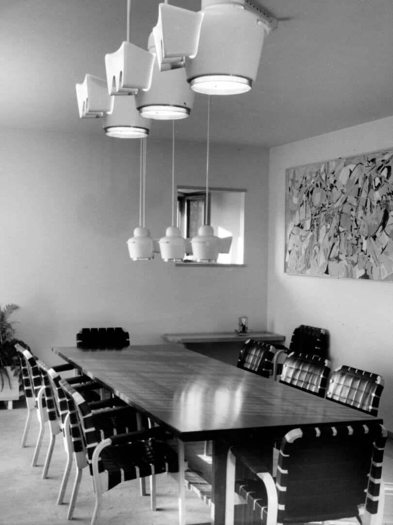 A modern dining room with a long wooden table, eight chairs with striped cushions, a contemporary light fixture, a piece of abstract art on the wall, and a small window in the background