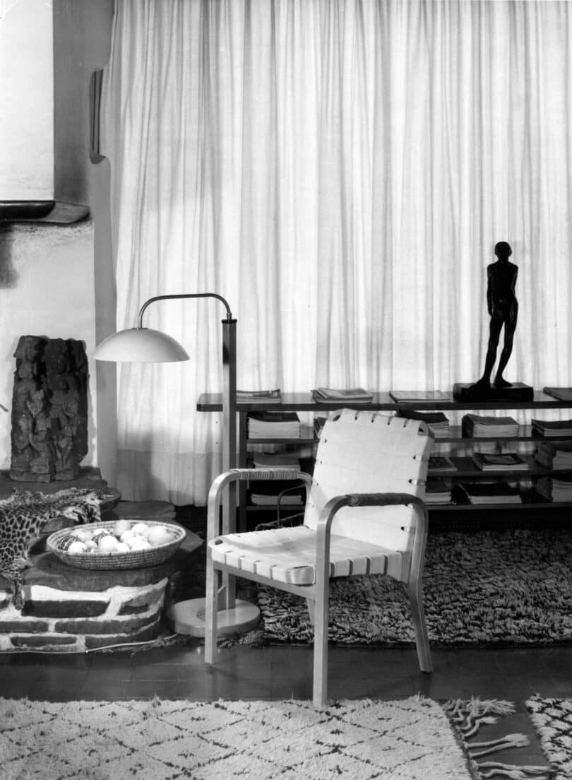 A modern living room features a white woven chair, a floor lamp, a statue, a bowl with stones, and assorted decor items on a rug, all set against a background of sheer curtains