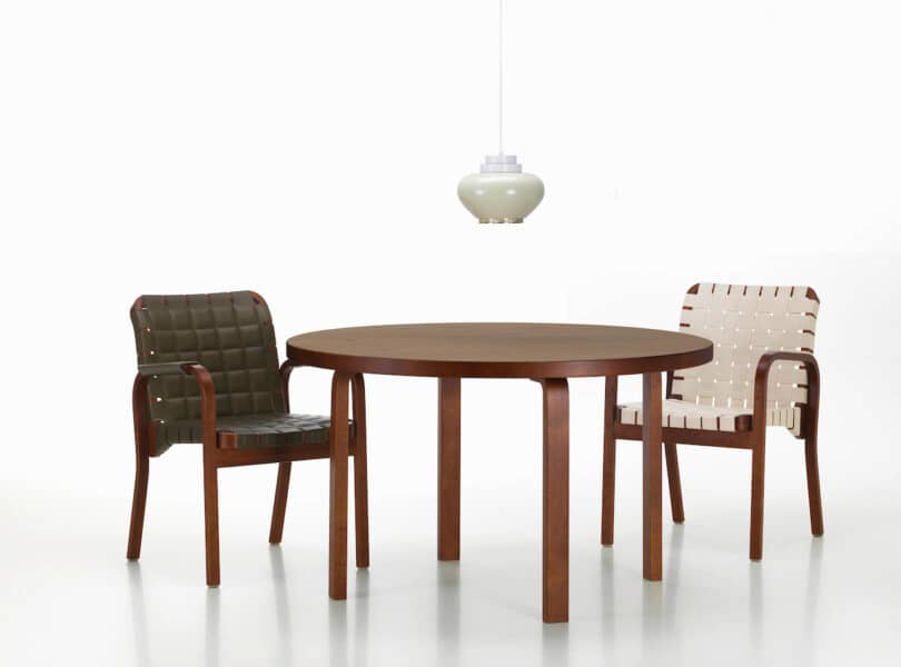 A round wooden table is flanked by two chairs with woven seats and backs; one chair is green and the other is white. A pendant light hangs above the table against a white background