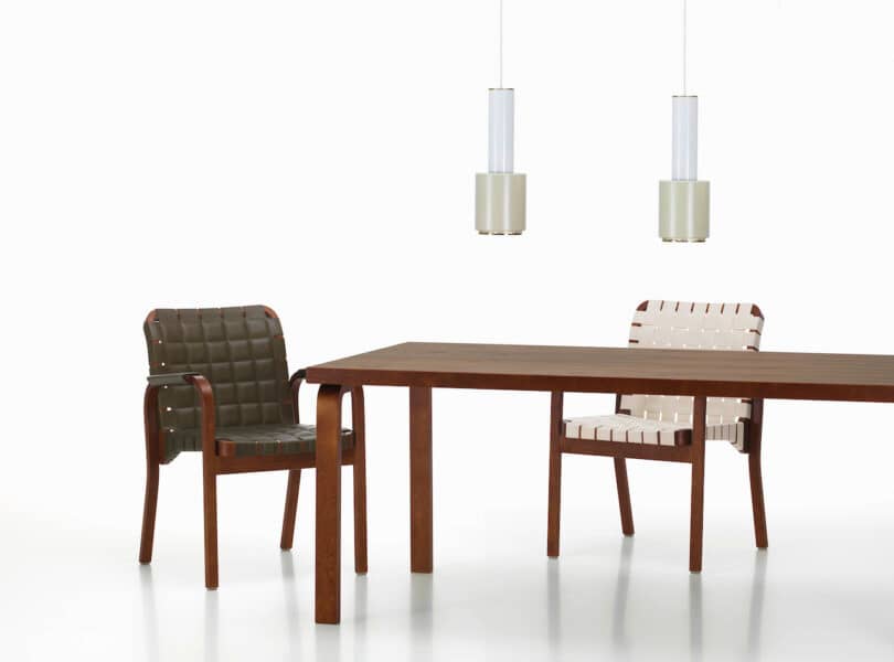 Two woven leather chairs, one green and one white, are positioned around a dark wooden table. Above the table are two modern pendant lights with cylindrical shades. The background is plain white