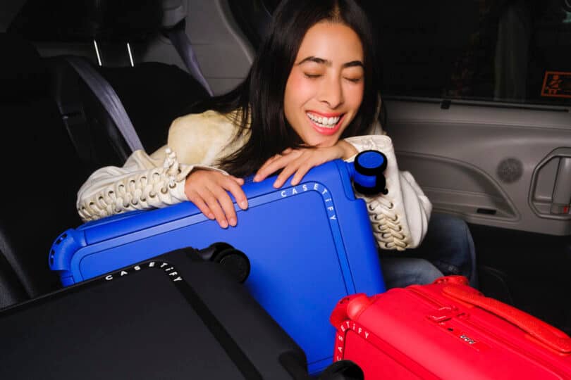 A person smiling while sitting in a car with blue, black, and red suitcases.