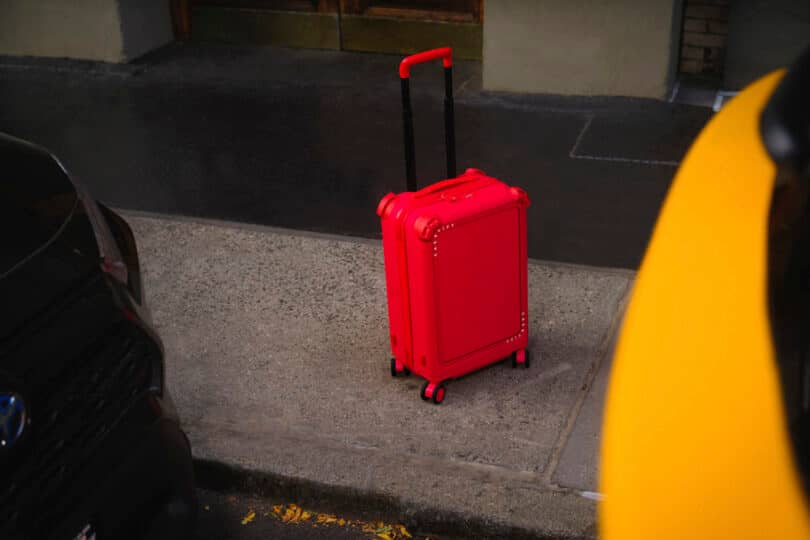 A red suitcase stands on a sidewalk between two parked vehicles.