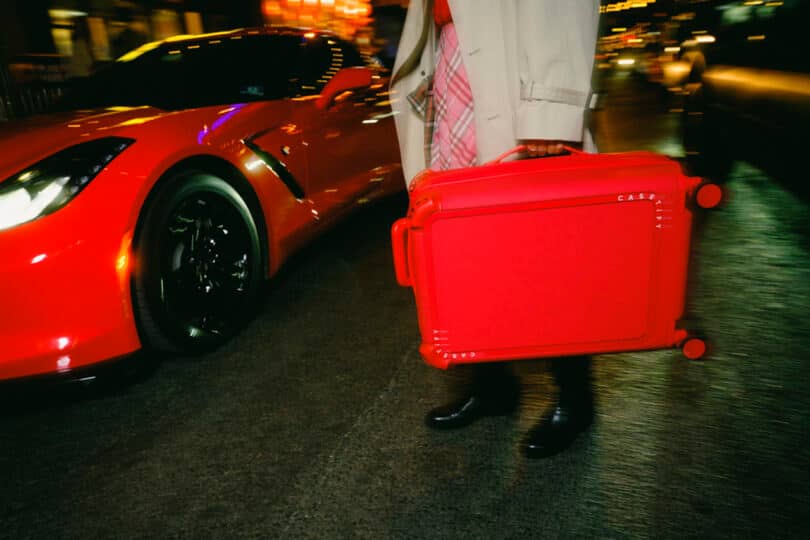 Person with a red suitcase walking beside a red sports car on a city street at night.