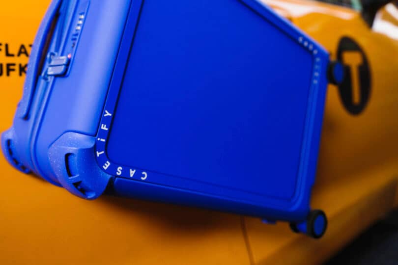 A blue suitcase is attached to the side of a yellow taxi with its wheels facing outward.