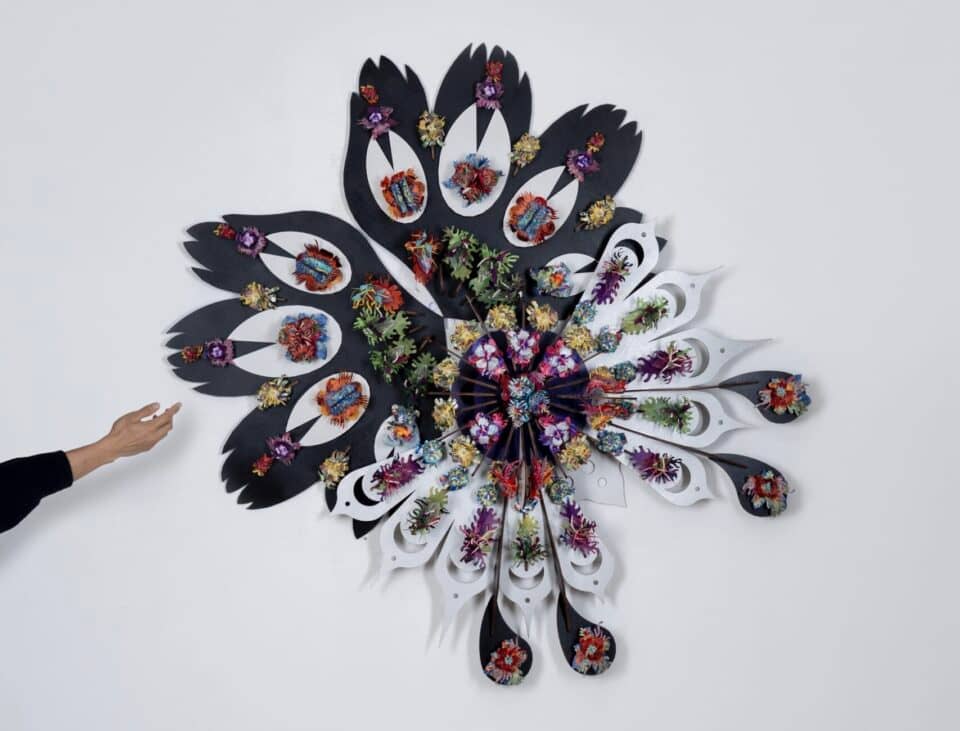 a sculptural wall piece made from mixed media, slightly resembling feathers that splay out from a central point, with a hand held up next to it for scale