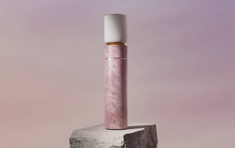 A tall, cylindrical object with a pink base and a white top stands on a rock pedestal against a soft, blurred pink and purple background.