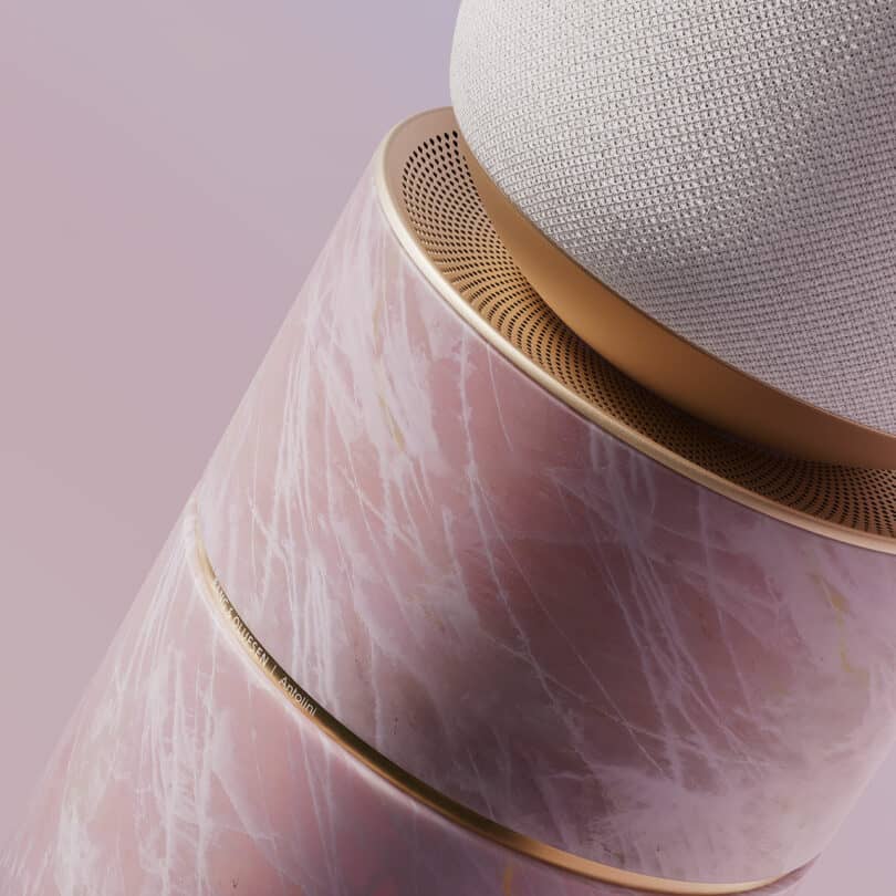 A round, textured, and fabric-covered speaker sits on a cylindrical pink and gold-marble base on a light pink background.